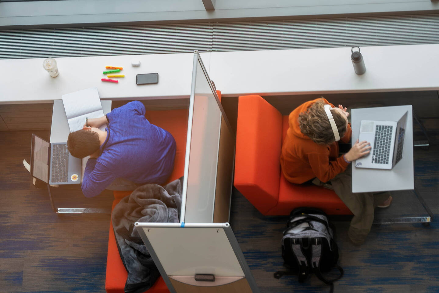 Two individuals study in separate nook areas of a modern library, one writing in a notebook and the other using a laptop with headphones on.